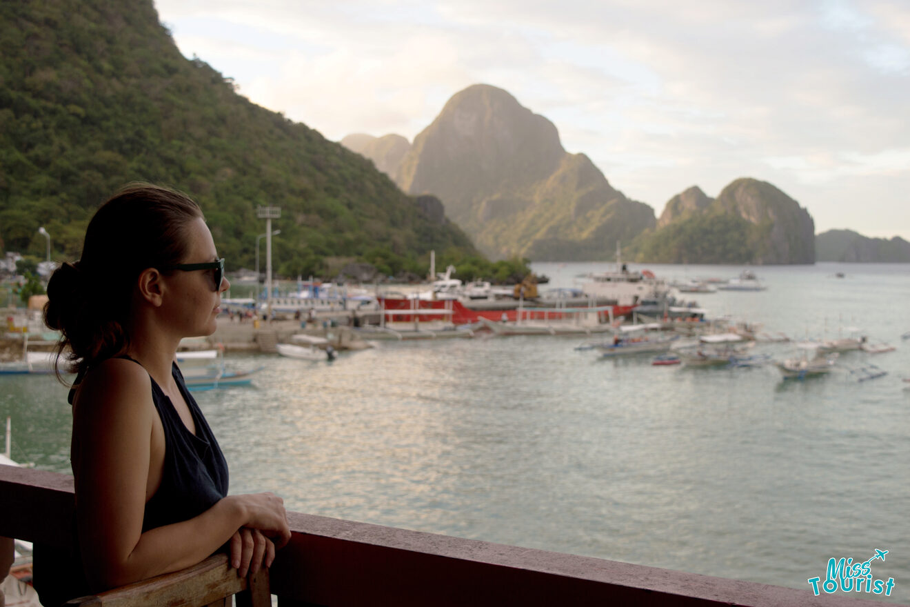 The writer of the post wearing sunglasses gazes out from a balcony at a scenic harbor filled with boats, framed by large limestone formations in the background.